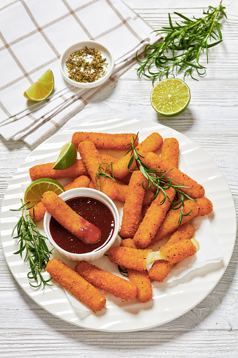 fried breaded mozzarella sticks served with dipping sauce, lime slices and fresh rosemary on white plate on white wooden table with ingredients, vertical view