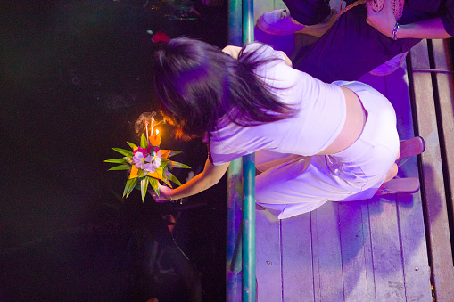 Young thai woman is placing   flower bouquet  onto water at  Loi Krathong festival. Woman has natural long black hair and is kneeling down  at local festival at canal in Bangkok Ladprao near Wanghin Rd. She is  opposite from temple Wat Siri Kamala Vas (Wat Senanikom).  View from footbridge over canal. Another person is close partially visible in frame