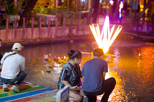 Thai couple is celebrating Loi Krathong at small pond at shopping and bar area Plaza Lagoon in Bangkok Ladprao at Wanghin Road.  Adult couple is praying. Another person is at  their left side. In background are neon lights on pond and flower bouquets are swimming on surface.