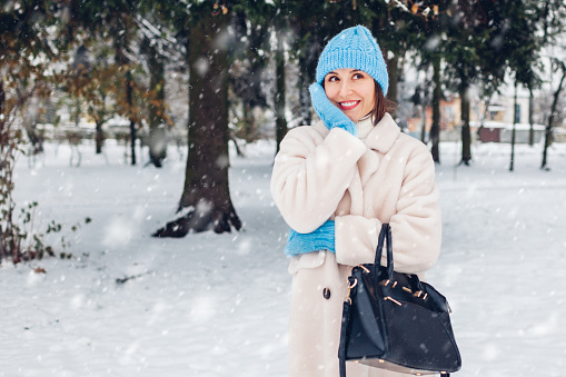 Happy smiling woman walking in snowy winter park wearing white sheepskin fur coat holding handbag enjoying landscape during snowfall. Warm clothes. Space