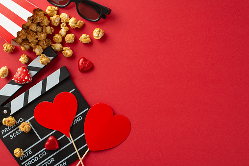 Top view composition of clapperboard, 3D glasses, popcorn, spilled from box, candies, and heart-shaped decor on a romantic red backdrop. Ideal for capturing the essence of love and cinematic charm