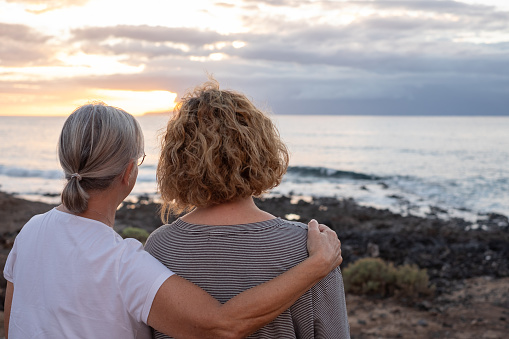 Back view of mature and senior women standing face to the sea looking at horizon over water, two bonding people embraced stay together enjoying freedom and vacation or retirement