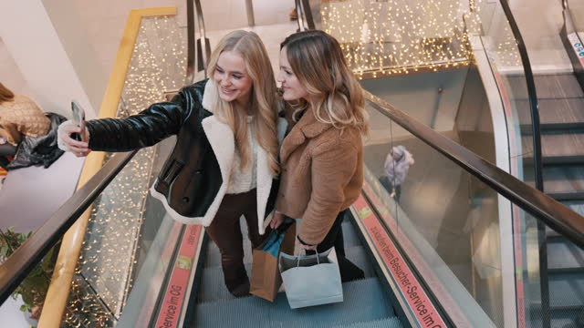 SLO MO Happy Young Female Friends in Trendy Clothes With Shopping Bags Taking Selfie on Escalator in Shopping Mall