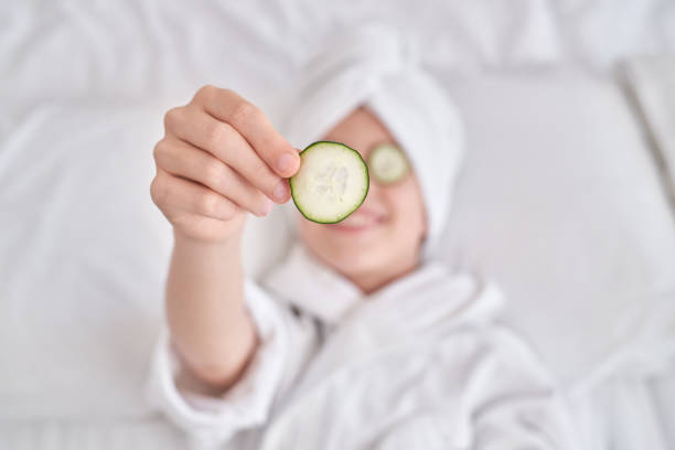 From above of child wearing bathrobe and towel turban lying on white bed and demonstrating cucumber slice during spa procedures in bedroom at home