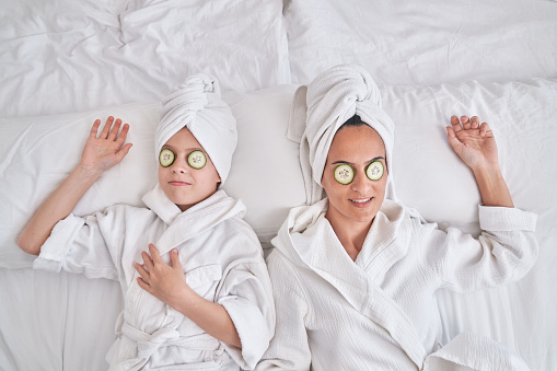 From above of relaxed woman and son in bathrobes with fresh cucumber slices on eyes lying together on bed during skincare routine