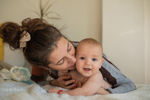 Adorable smiling baby boy is lying on his tummy on the white blanket on the bed and looking at camera while his mother is above him and hugging and kissing him. They are enjoying the moment. Simple happy life.