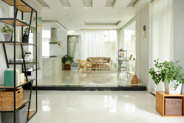 The interior of a home, with the dining room in the middle, the living room to the right, the kitchen behind the dining room and a hallway to the left.  The floor is multicolored wood, and the furniture is modern.  In the foreground of the image is a deck to an outside area.