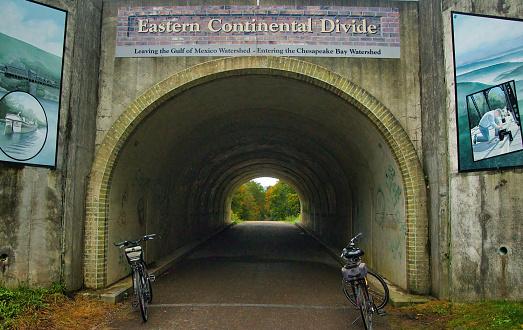On a cloudy day in early Autumn, two bikes are parked beside a tunnel with signs indicating the Eastern Continental Divide along the Great Allegheny Passage recreation trail.
