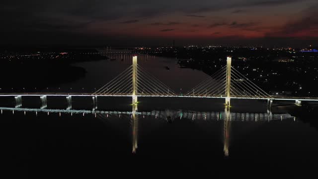 A cable-stayed highway bridge with tall concrete pylons illuminated by architectural lighting at night. A panorama of the riverbed running away under the night sky against the background of city lights