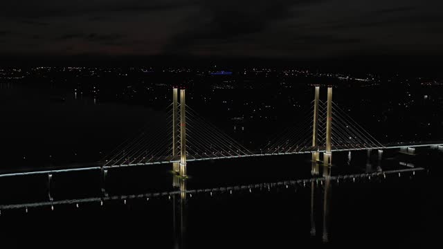 A cable-stayed highway bridge with tall concrete pylons illuminated by architectural lighting at night. General plan of the bridge against the background of night city lights, flying backward