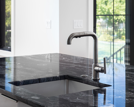 A modern kitchen black stainless faucet detail on an island with a black marble countertop, black frame windows, and white walls.