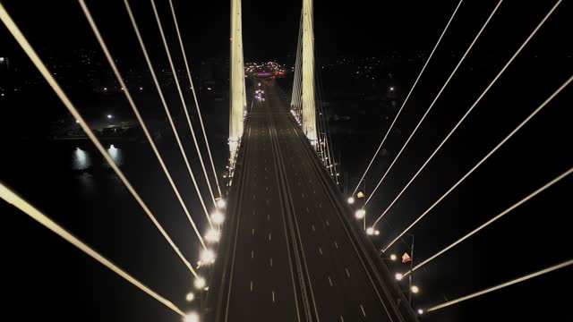 A cable-stayed highway bridge with tall concrete pylons illuminated by architectural lighting at night. Slow span between the bridge piers, motorcycles and cars driving below