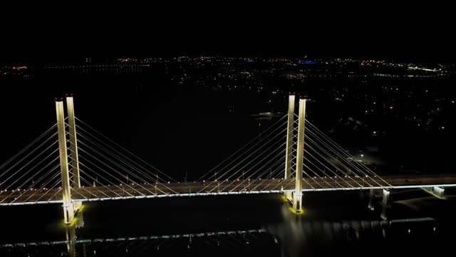 A cable-stayed highway bridge with tall concrete pylons illuminated by architectural lighting at night. Fly back, opening view of the bridge, city illumination in the background