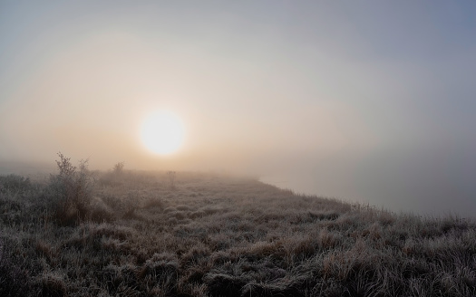 Soft focus. The sun rises over the field in a thick fog. Minimalistic natural foggy landscape. The shore of the lake is barely visible in the morning fog. Panoramic view.