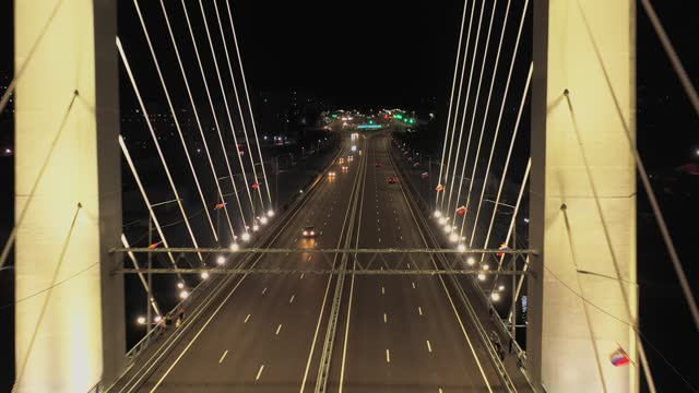 A cable-stayed highway bridge with tall concrete pylons illuminated by architectural lighting at night. Flying over the bridge roadbed between the pylons, automobile traffic below