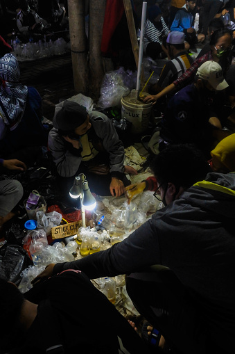 As the azure hue of the Indonesian sky shifts into a tapestry of twilight, a vibrant scene emerges at the Jatinegara night market. Here, an intimate circle forms—a fusion of silhouettes against the soft backdrop of market life. This image captures a spontaneous congregation of people, grounded upon the earth, bathed in the warm, dim light that trickles down from strings of bulbs above. They gather not merely for transactions but for the camaraderie that thrives amid the exotic aquatic bazaar. Their postures relaxed, their attention tuned to the freshness of the day's catch, presented in woven baskets and makeshift stands, this group embodies the spirit of the market: collective, authentic, alive. Beyond a mere marketplace, this photograph encapsulates a community's heart, pulsing in harmony with the nocturnal rhythm of Jatinegara