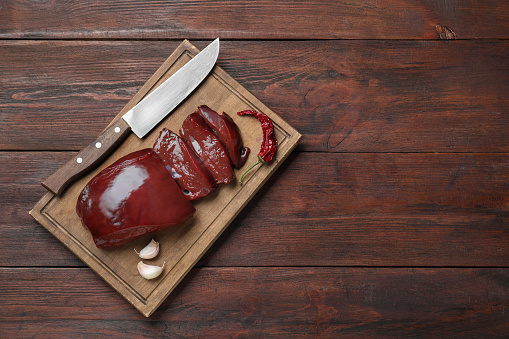 Cut raw beef liver with chili pepper, garlic and knife on wooden table, top view. Space for text