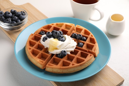 Tasty Belgian waffle with blueberries, honey and whipped cream on white table, closeup