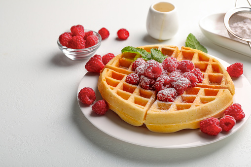 Tasty Belgian waffle with fresh raspberries, powdered sugar and mint on white table, space for text