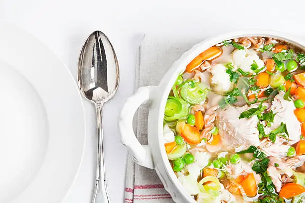 bowl filled with healthy chicken soup, made of chicken, whole wheat pasta, and lots of vegetables, on white tablecloth