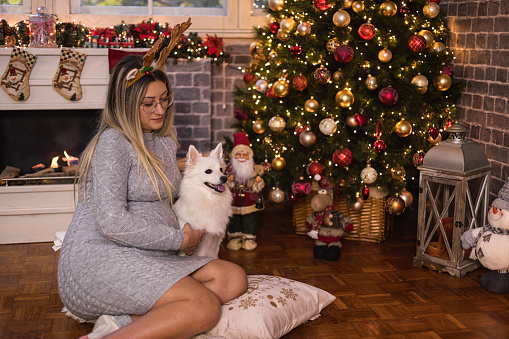 Young beautiful pregnant woman sitting on a floor and playing with her dog in a cozy Christmas atmosphere