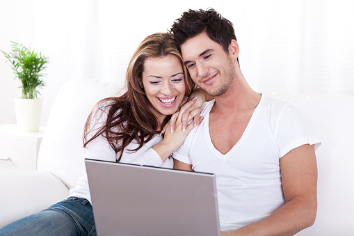 Smiling young couple sitting on white sofa working on laptop computer.