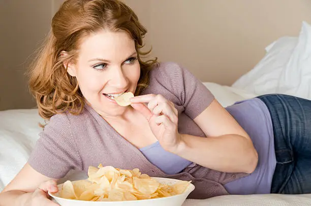 Woman with a bowl of chips lying on a bed.