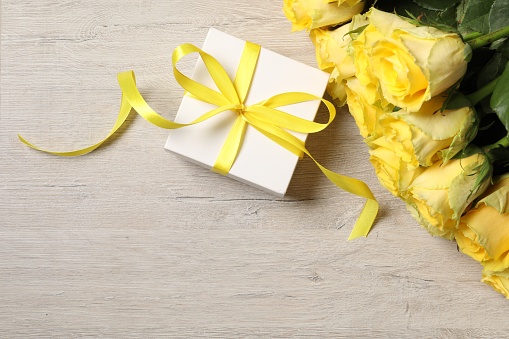 Beautiful bouquet of yellow roses and gift box on wooden table, flat lay. Space for text