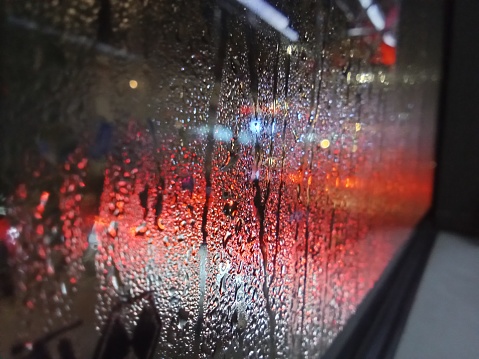 Abstract image depicting glowing city lights - coming from traffic and storefronts - defocused beyond a raindrop-soaked window.