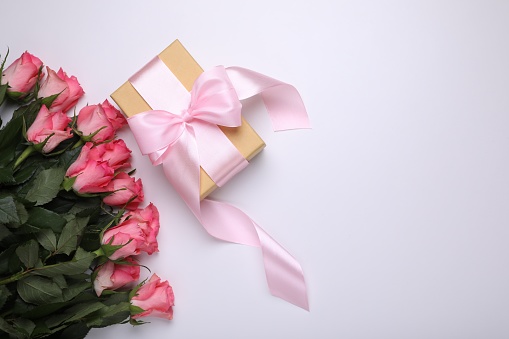 Gift box wrapped with pink bow and beautiful flowers on white background, flat lay. Space for text