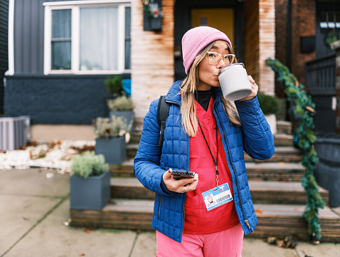 Asian female registered nurse going to work in the morning and drinking morning coffee. She is wearing eyeglasses, medical scrubs and winter jacket and hat. Exterior of urban home in Hamilton, Canada.