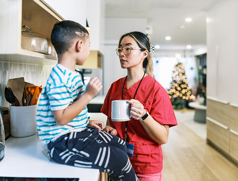 Asian female registered nurse and her young son getting ready for the day. She is wearing eyeglasses and pink  medical scrubs, boy is wearing casual clothes. Kitchen interior of urban home in Hamilton, Canada.