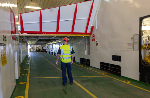 12th May 2023: View of the parking deck of a Caledonian MacBrayne ferry, leaving Uig on the Isle of Skye.  A crew member in high visibility workwear is preparing to guide vehicles aboard, having had the hydraulic level above raised with cars already securely loaded. The port is a busy ferry terminal that connects the Highlands and islands on the northwest coast of Scotland.
