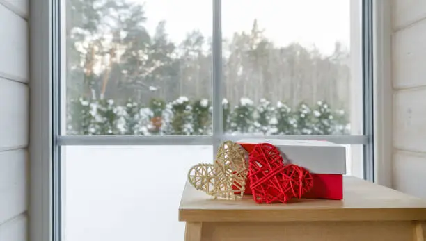 A gift in a red-white box and two hearts on a wooden table near the window with a view of the winter garden, Scandinavian style. Valentine's day concept