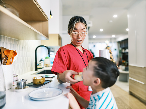 Asian female registered nurse and her young son getting ready for the day. She is wearing eyeglasses and pink  medical scrubs, boy is wearing casual clothes. Kitchen interior of urban home in Hamilton, Canada.