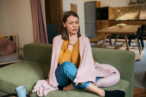Caucasian woman suffering from sore throat sitting on sofa. Sick young woman covered with blanket coughing while sitting on the sofa at home.