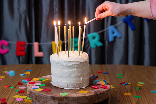 A person in a pink, long sleeve lights tall decorative candles of white and gold; on a cake with white frosting, that is taller than it is wide. There is a steel blue curtain background with the word 'celebrate' draped across. The cake stand and table the cake are on are both made of wood. The table has circular, paper confetti thrown on it.