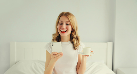 Happy smiling young woman with mobile phone holding cup of coffee in bed in white bedroom at home in the morning