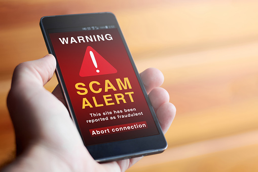 Closeup of male hand holding modern mobile phone with red screen and warning symbol with the text SCAM ALERT: This site has been reported as fraudulent. Abort connection. Concept of internet security.