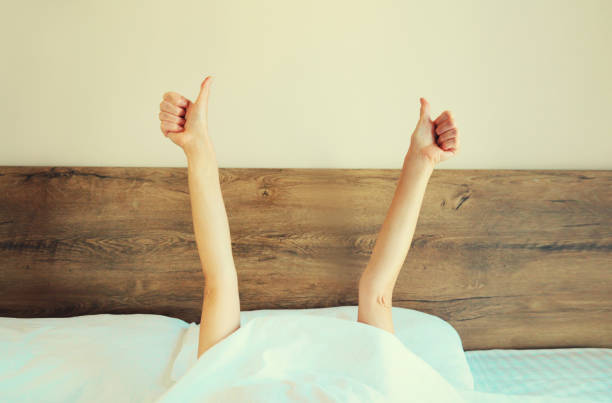 istock Cheerful lazy woman waking up after sleeping lying in soft comfortable bed showing gesture stretching her hands up from under the blanket in white bedroom 1836008288