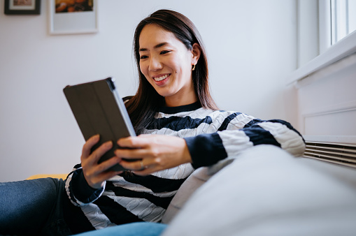 Happy young woman spending time at home sitting on a couch and browsing news on a digital tablet