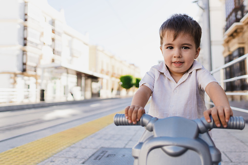 Portrait of little boy in the street looking at camera while sitting on his blue toy motorcycle.
