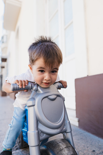 Little boy is standing with his ride-on in the middle of the street.