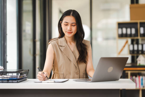 Asian freelance writer takes notes at her desk with a laptop in a bright, modern office setting