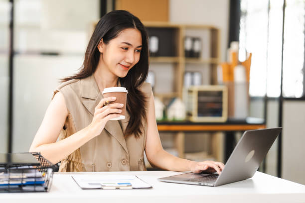 Asian freelance writer takes notes at her desk with a laptop in a bright, modern office setting