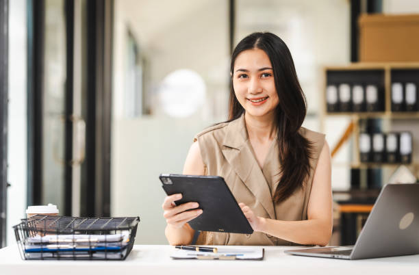 Asian freelance writer takes notes at her desk with a laptop in a bright, modern office setting