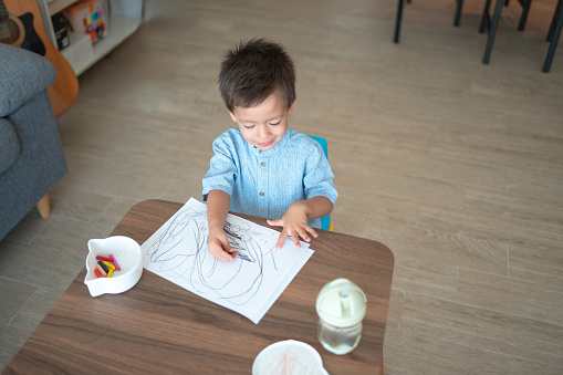 In the cozy living room, cute and multiracial two and a half year old boy siting on a small chair, engrossed in a world of imagination. With sheer delight, he draws vibrant strokes with crayons on a white paper, laid out on a small wooden table. Heartwarming scene of early artistic exploration
