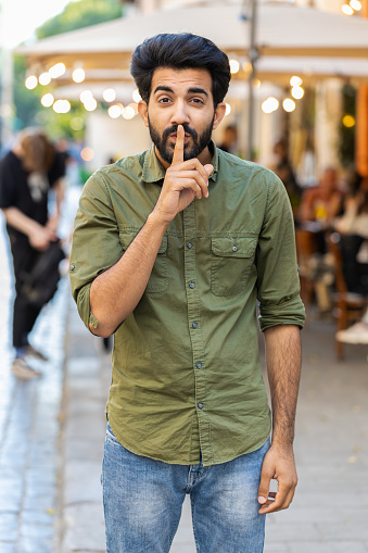 Shh be quiet please. Indian man presses index finger to lips makes silence hush gesture sign do not tells gossip secretoutdoors. Arabian guy walking in urban city street. Town lifestyles. Vertical