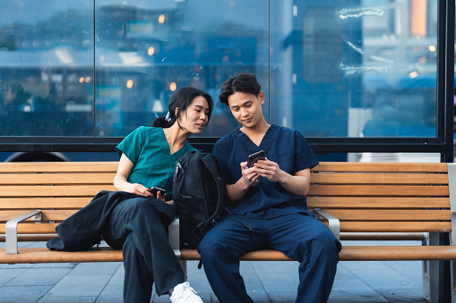 Two nurses in the medical scrubs sitting waiting for the bus at bus station using smartphone.