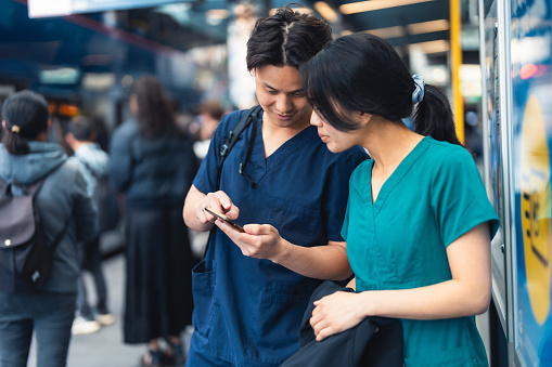 Nurses in medical scrubs outdoor waiting for the bus using smartphone.